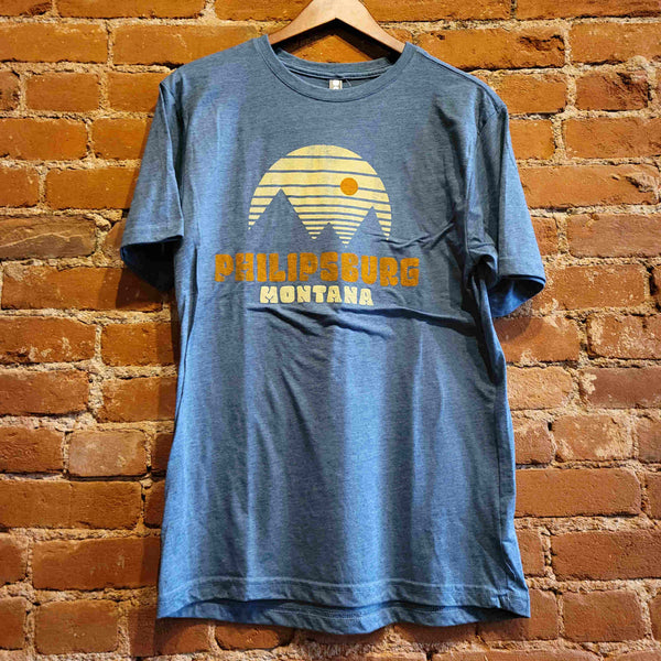 Front of men's crew neck t-shirt. The shirt is a blue jean color with the text "Philipsburg Montana" printed in burnt orange and cream colored ink. Above the text is an  outline of pointy mountains under a striped cream colored sky with an orange sun. 