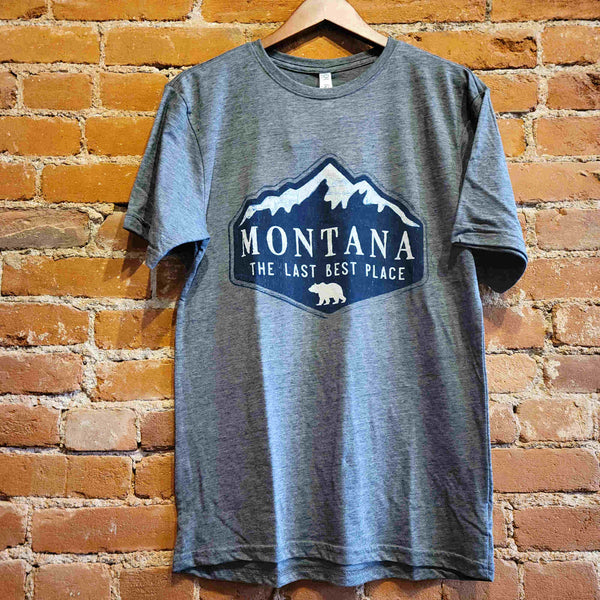Front of the men's crew neck t-shirt. "Montana The Last Best Place" is printed in white ink surrounded by dark blue snow capped mountains. Below the text is the silhouette of a grizzly bear printed in white ink. 