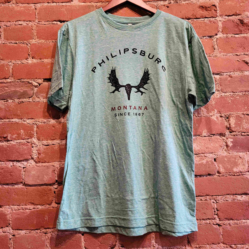 Front of the Men's crew neck short sleeved shirt. There is a moose skull printed in black along with the text 'Philipsburg Montana Since 1867". Montana is printed in red, The t shirt is a bright jade color with little white tweed specs throughout the fabric. 