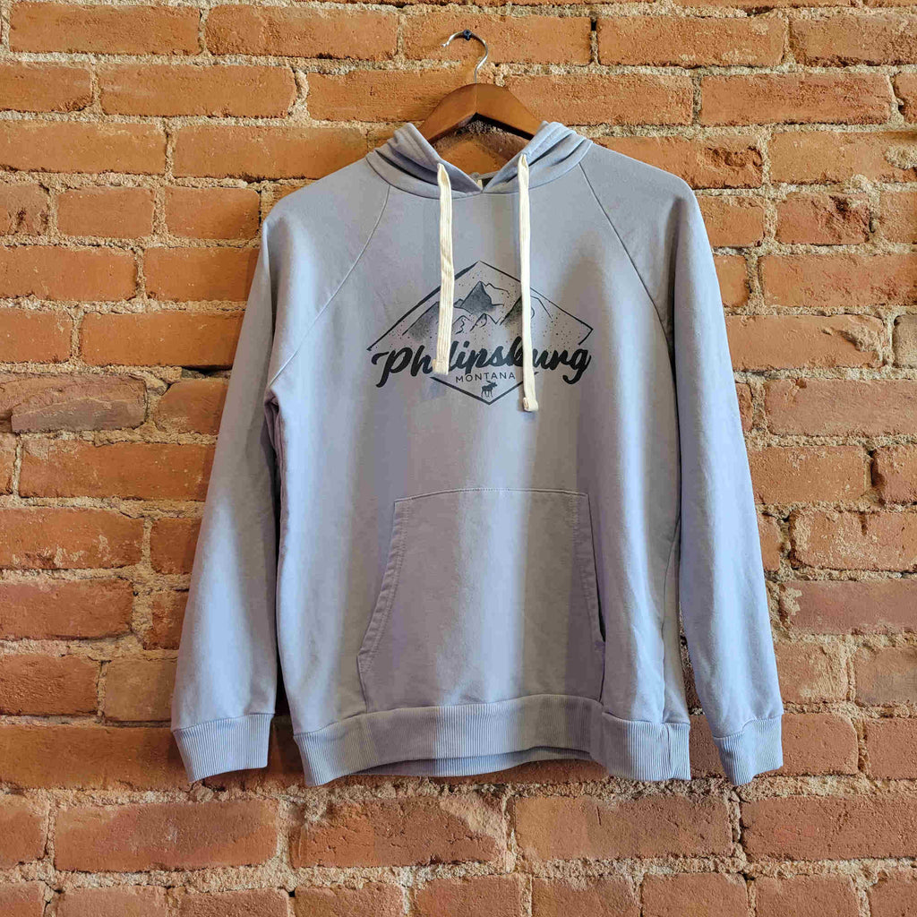 Front view of the women's mineral wash hoody. The hoody is light blue with a dark blue print on the front. The print features mountains inside a diamond silhouette. "Philipsburg Montana" is placed below the mountains along with a small moose sillouete . There are white drawstrings, and a kangaroo pocket.