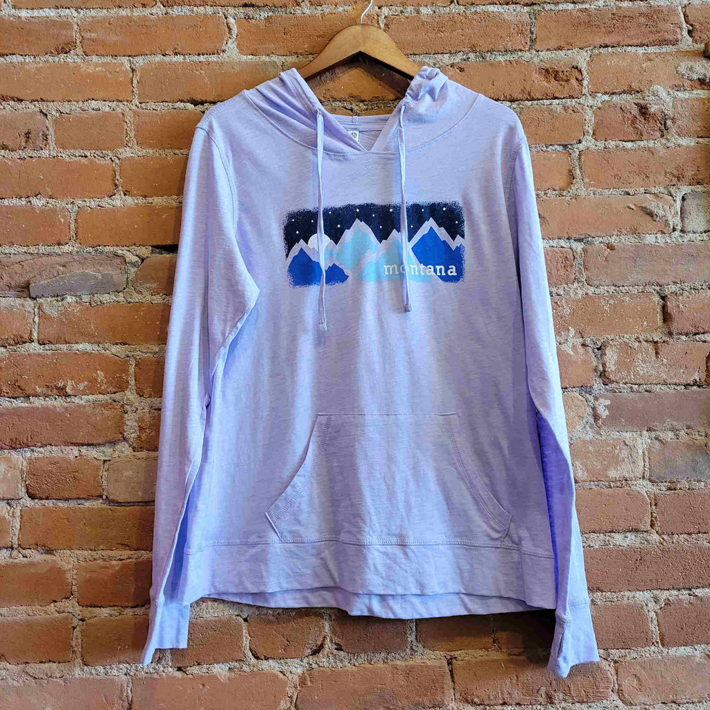 Picture of Ouray women's lightweight hoody in the colorway pastel lilac (light  purple). The front features jersey drawstrings, thumbholes, and a kangaroo pocket.  The print depicts a blue mountain range with a starry night sky. "Montana" is printed on the bottom right corner of the image.