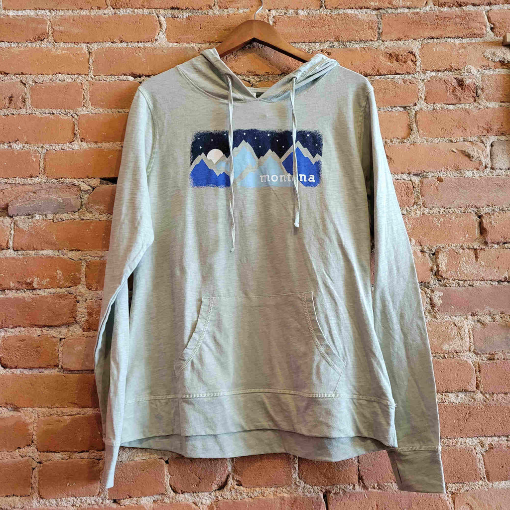 Picture of Ouray women's lightweight hoody in the colorway Desert Sage (light olive green). The front features jersey drawstrings, thumbholes, and a kangaroo pocket.  The print depicts a blue mountain range with a starry night sky. "Montana" is printed on the bottom right corner of the image.
